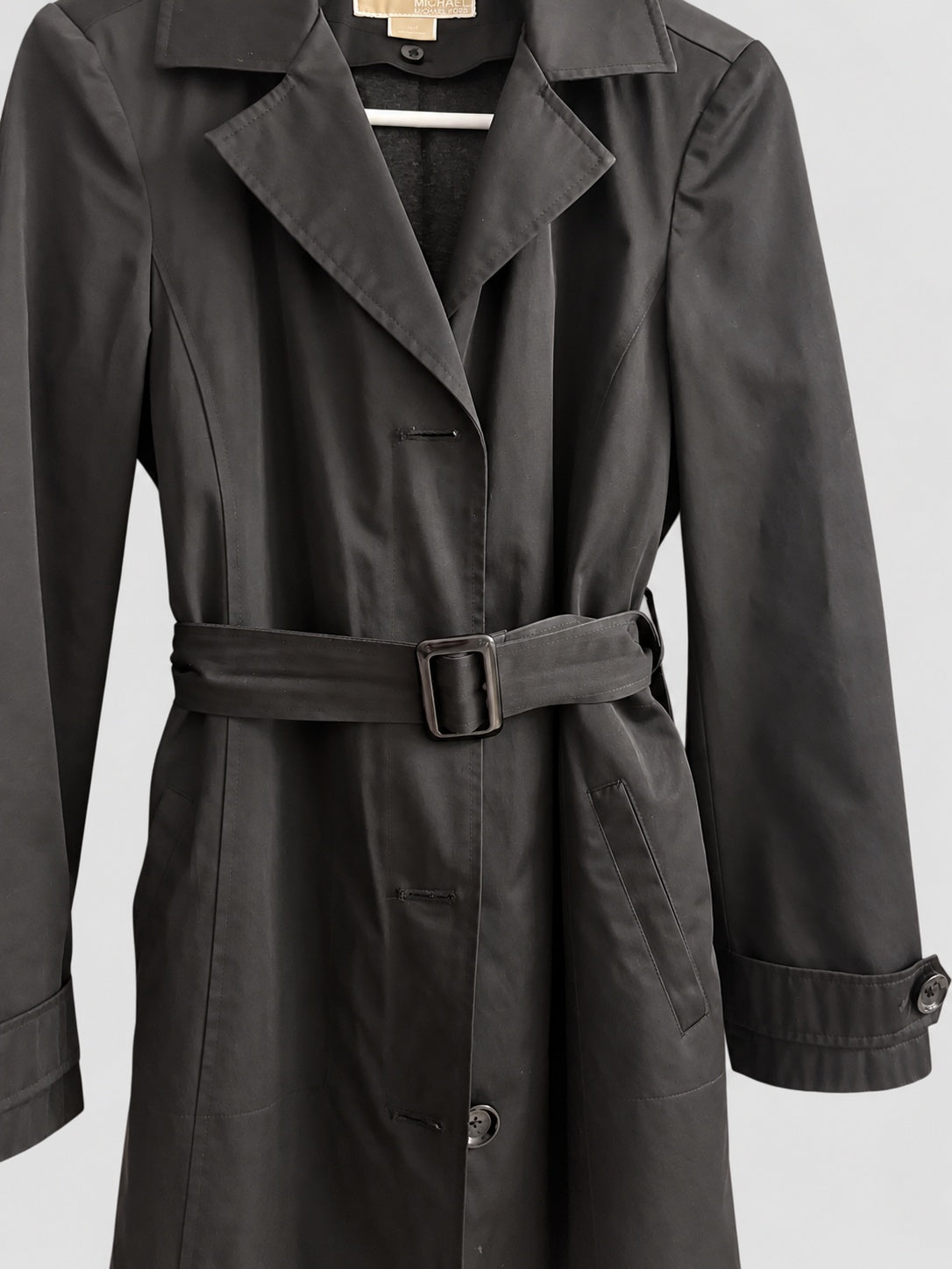 A black trench coat with a matching waist belt and buckle, shown on a hanger against a light gray background.