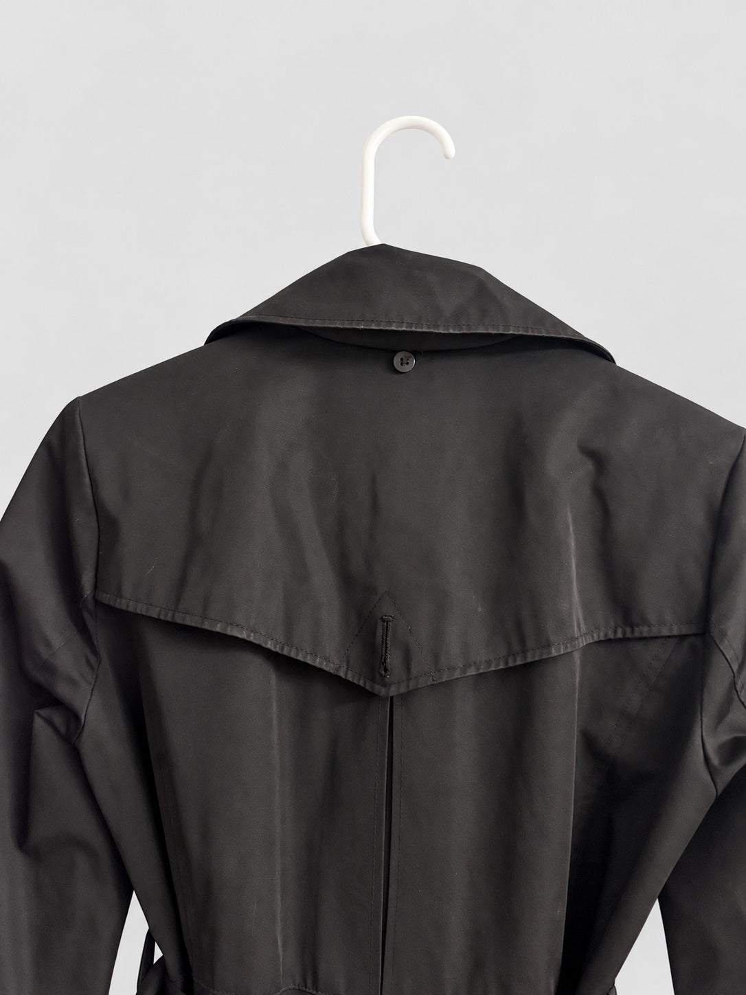 A black trench coat hanging on a white plastic hanger against a light gray background, showing the back view with a button closure and shoulder flap.