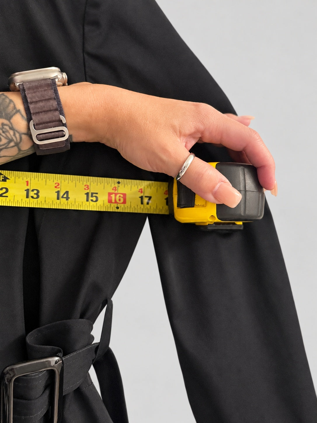 A close-up of a person's hand holding a yellow tape measure against the sleeve of a black belted coat or dress, showing garment measurements.