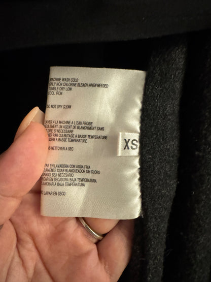 A close-up of a hand holding a white garment care label inside a black garment. The label shows care instructions and a size marking of XS.