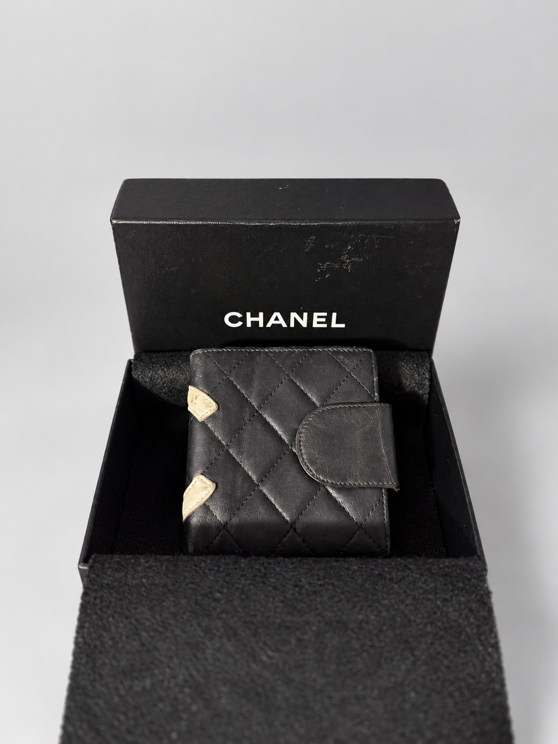 A black quilted leather wallet with gold-tone corner accents, presented inside an open black Chanel box on a light gray background.