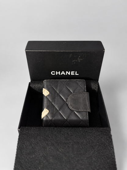 A black quilted leather wallet with gold-tone corner accents, presented inside an open black Chanel box on a light gray background.