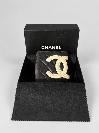 A Chanel black quilted wallet with a gold-tone interlocking CC logo, presented inside its black Chanel box with a black microfiber pouch on a light gray background.