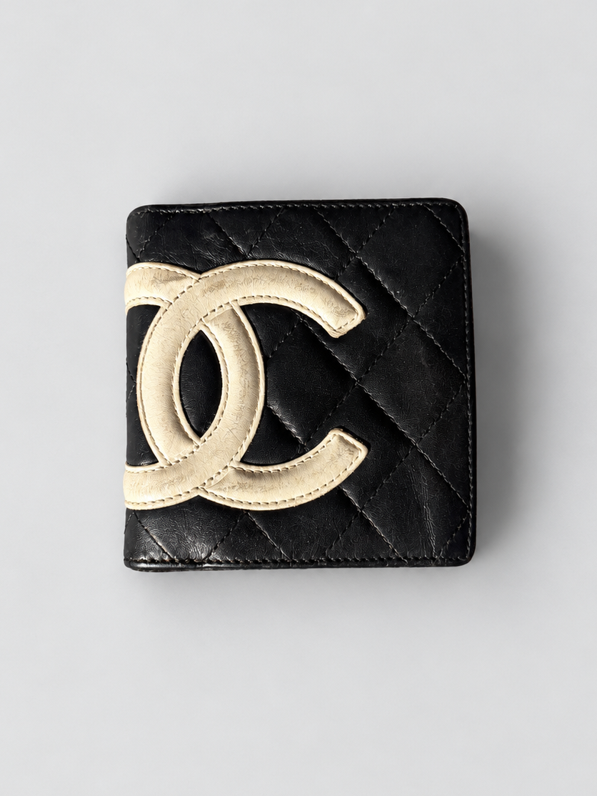 A black quilted leather Chanel bi-fold wallet featuring a large, raised gold-tone interlocking CC logo on the front, set against a neutral light gray background.
