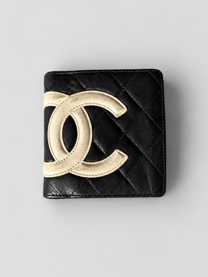 A black quilted leather Chanel bi-fold wallet featuring a large, raised gold-tone interlocking CC logo on the front, set against a neutral light gray background.