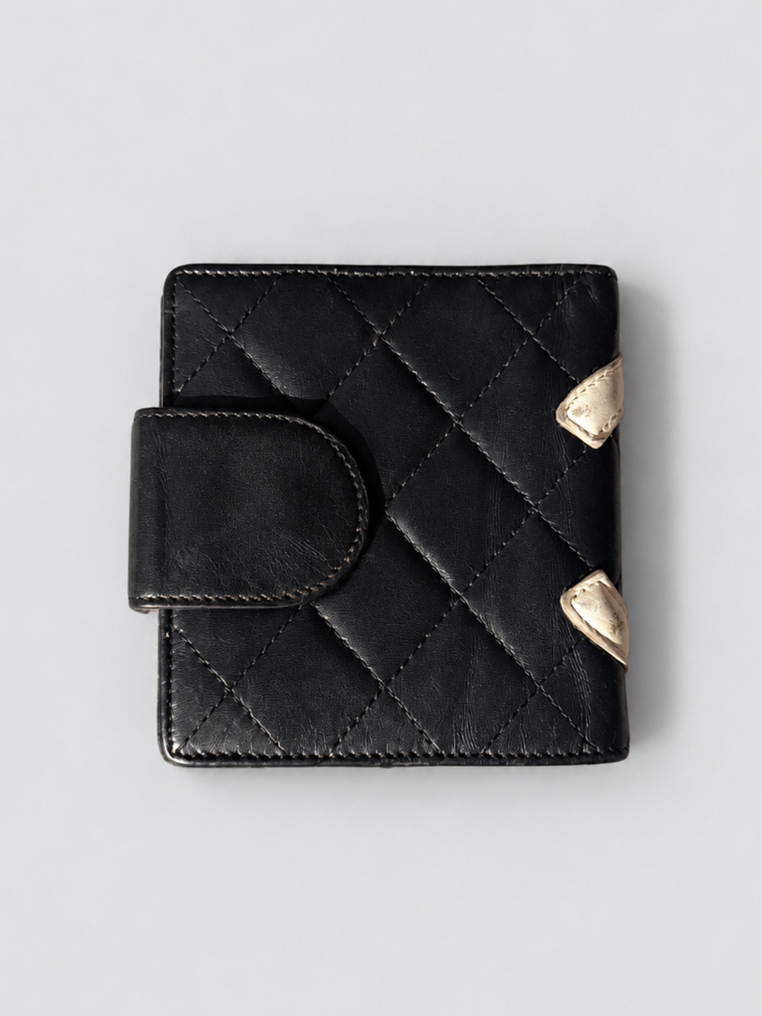 A black quilted leather wallet with a snap tab closure and two small gold-tone shield-shaped metal accents on the side, shown on a light gray background.