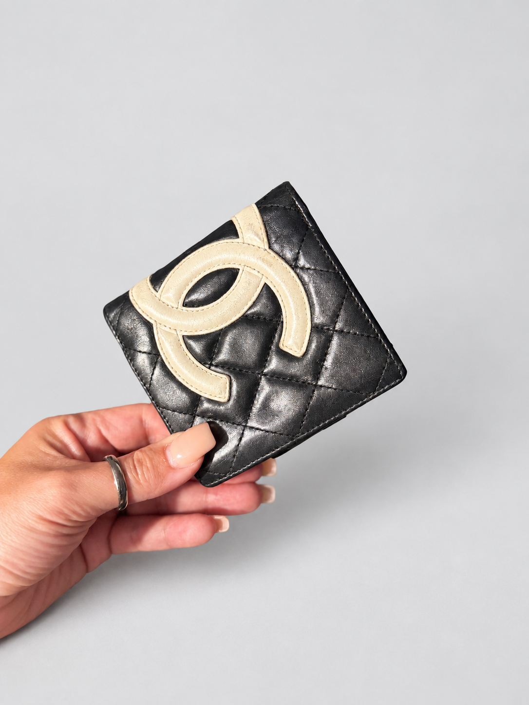 A hand holding a black quilted Chanel wallet with a large beige CC logo on the front, set against a plain light gray background.