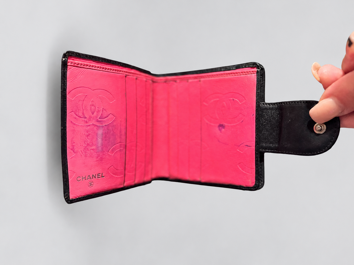 A Chanel bi-fold wallet in black leather with a vibrant pink interior, held open by a hand against a light gray background. The interior features an embossed Chanel logo and the brand name 'CHANEL' printed in the bottom right corner.