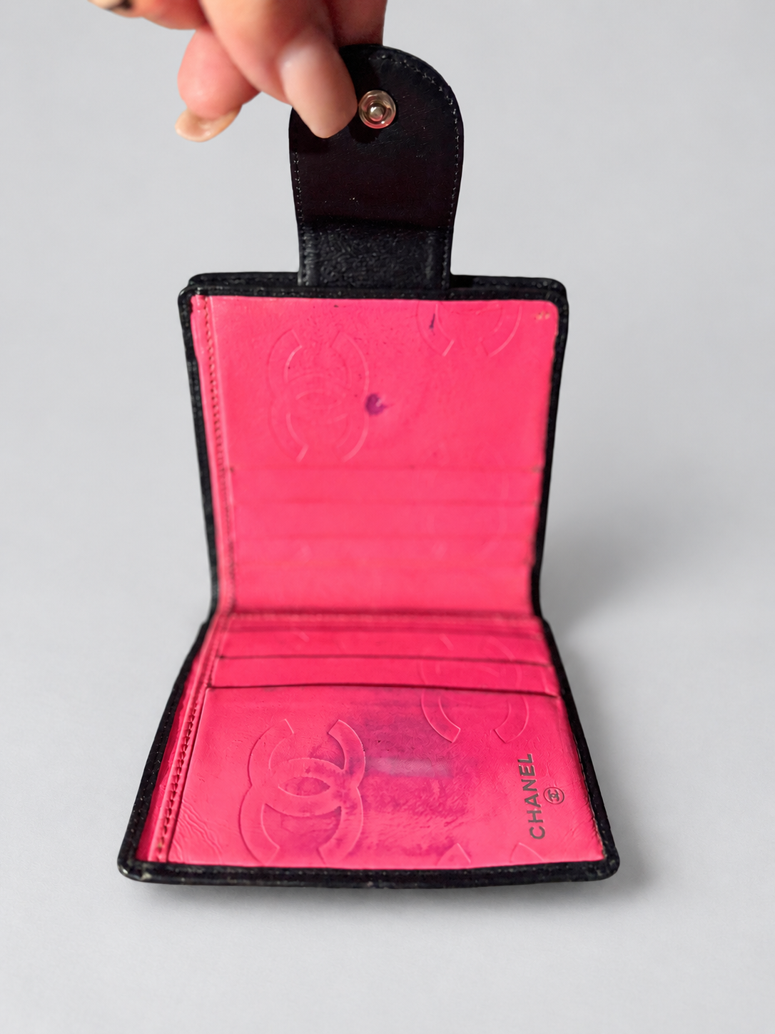 A Chanel bi-fold wallet in black leather with a vibrant hot pink interior, held open by a hand. The interior features embossed Chanel logo patterns and card slots.