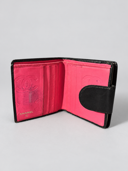 An open Chanel bi-fold wallet in black leather with a vibrant hot pink interior, featuring embossed Chanel logos and card slots.