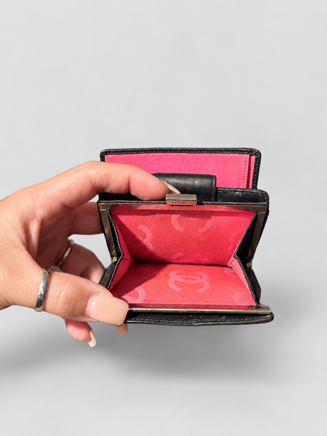A hand holding an open black leather clutch wallet with a bright pink interior featuring a subtle monogram pattern.