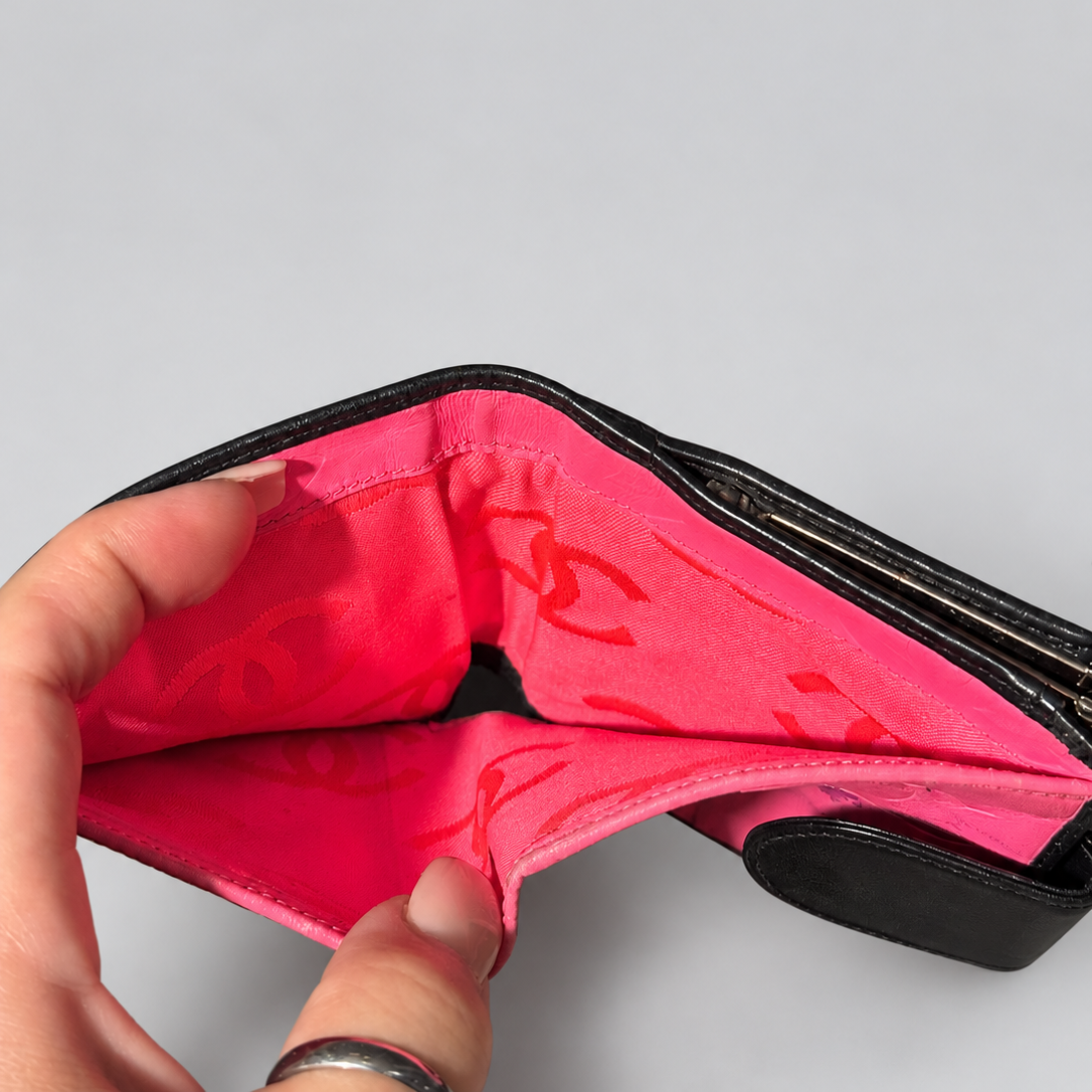 A close-up of a hand holding open a black wallet to show a bright pink interior with a repeating red monogram pattern, set against a plain light gray background.