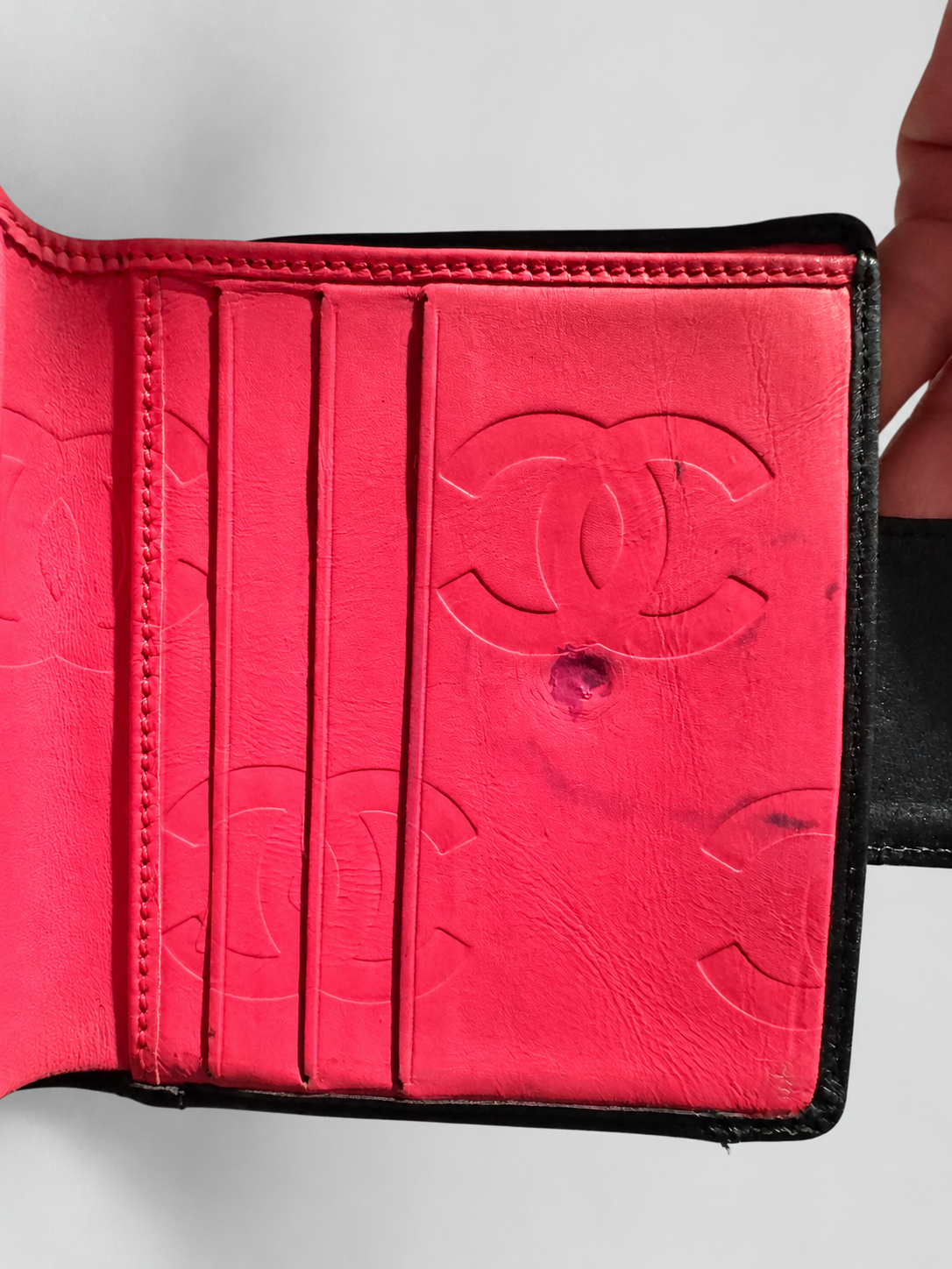 A close-up of an open Chanel wallet showing a bright pink leather interior with embossed Chanel logo pattern and card slots, held open by a hand against a light gray background.
