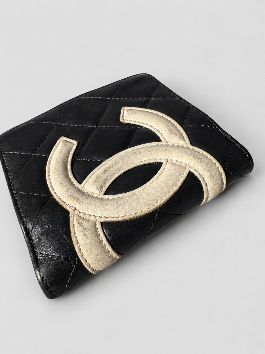 A black quilted leather wallet featuring a large, raised cream-colored interlocking CC logo on the front, characteristic of Chanel.