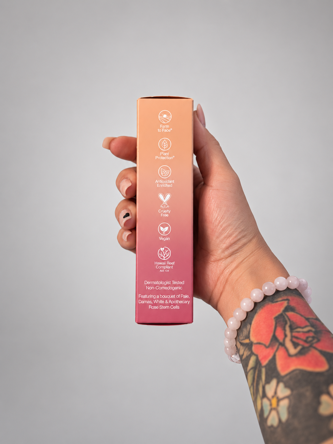 A hand with a pink beaded bracelet and a red floral tattoo holds a slim rectangular box with a peach-to-pink gradient. The box features white text and icons detailing the product's benefits, including 'Dermatologist Tested' and 'Vegan'.
