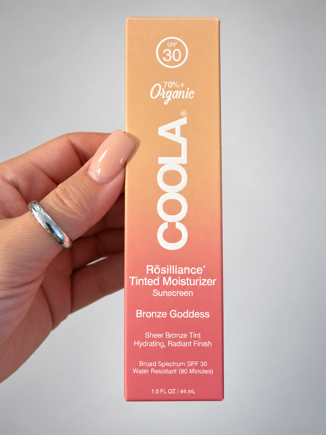 A hand with light pink manicured nails holds a tall rectangular box of COOLA Rósilliance Tinted Moisturizer Sunscreen in the shade Bronze Goddess. The box features a warm gradient from peach to pink, with white text.
