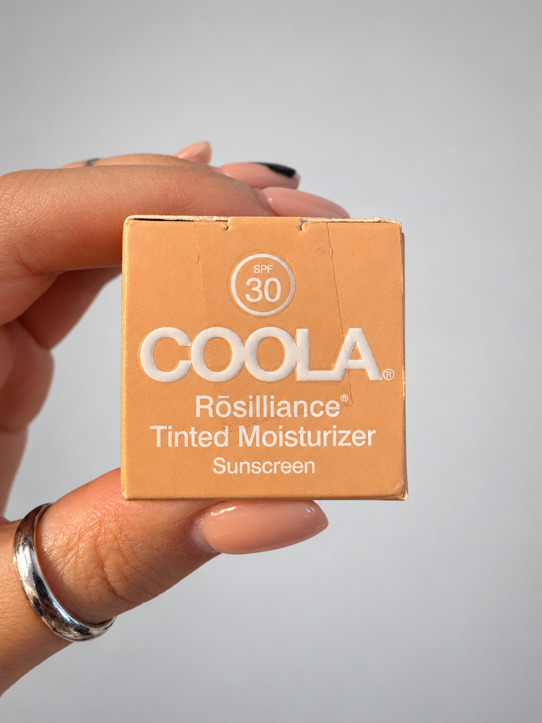 A hand holding a square cardboard box of COOLA Rósilliance Tinted Moisturizer Sunscreen SPF 30. The box is a warm peach-orange color with white text, held against a neutral light gray background.