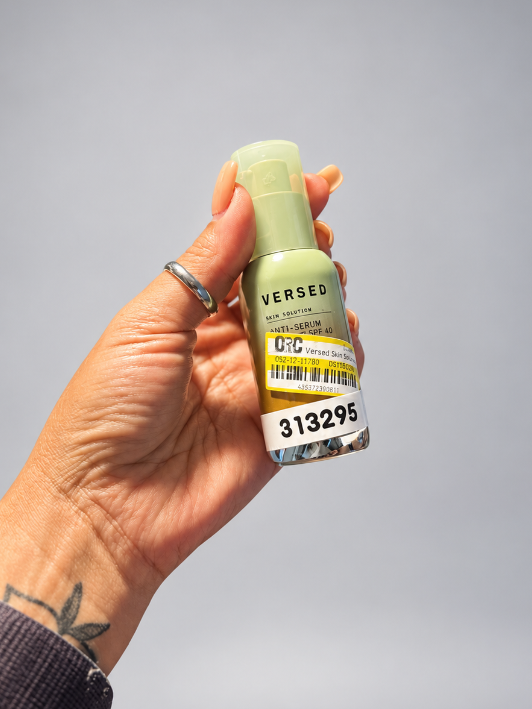 A hand holding a small bottle of Versed hair serum against a plain gray background. The bottle is a pale yellow-green color with a white and yellow barcode sticker on the front.
