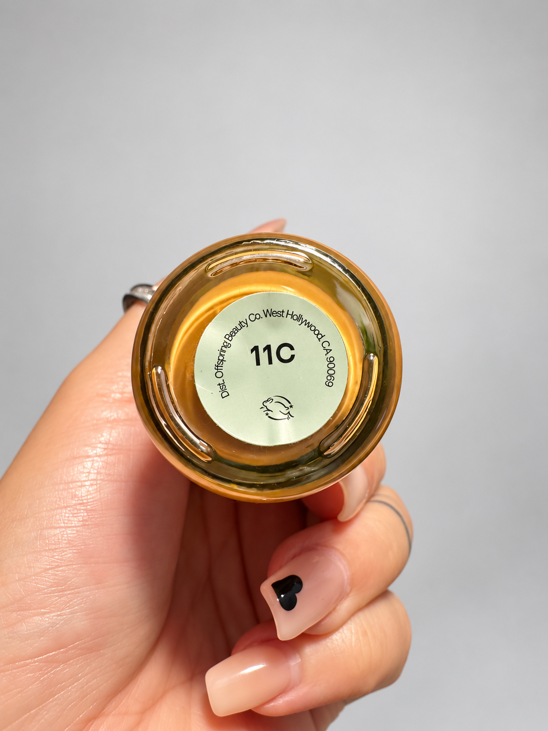 A hand holding the bottom of a gold cosmetic bottle, showing a circular label with the shade code '11C' and text reading 'Color Off-White Co. West Hollywood, CA 91111'.