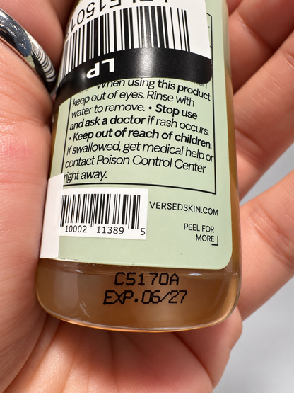 Close-up of the back of a Versed Skin liquid product bottle held in a hand, showing a barcode, warning label text, and amber-colored liquid inside a clear bottle.