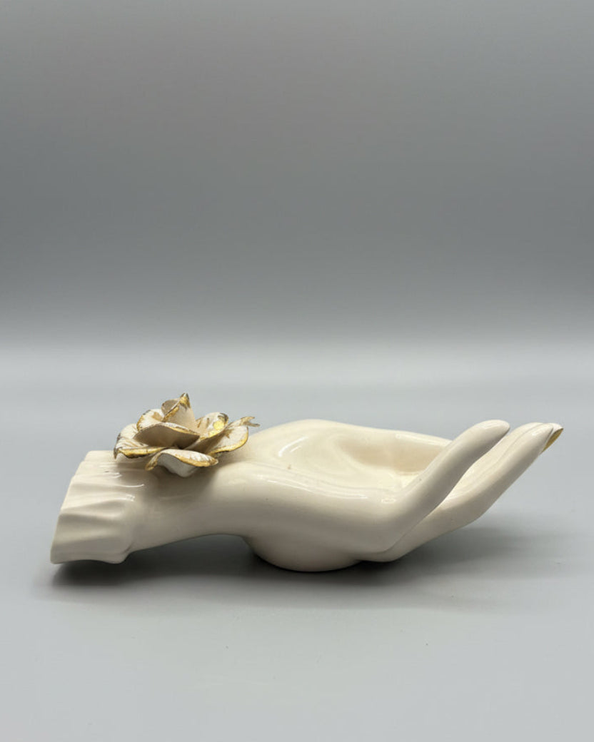 A cream-colored ceramic hand figurine holding a gold-toned flower, displayed on a dark wood tabletop with window blinds in the background.