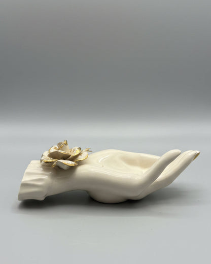 A cream-colored ceramic hand figurine holding a gold-toned flower, displayed on a dark wood tabletop with window blinds in the background.