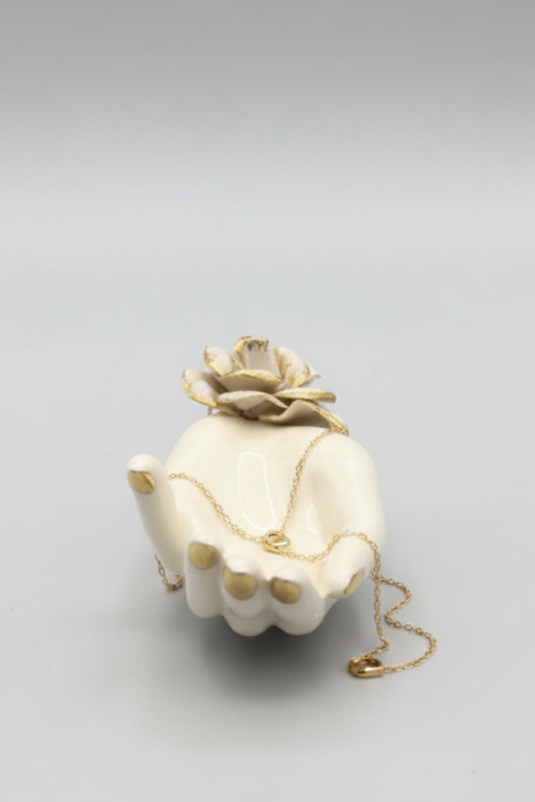 A small cream-colored ceramic figurine shaped like a hand holding a flower, with gold accents on the flower and fingers, placed on a dark wooden tabletop.