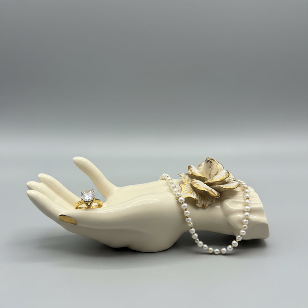 A cream-colored ceramic hand sculpture with a gold-toned flower accent, resting on a dark wood tabletop.