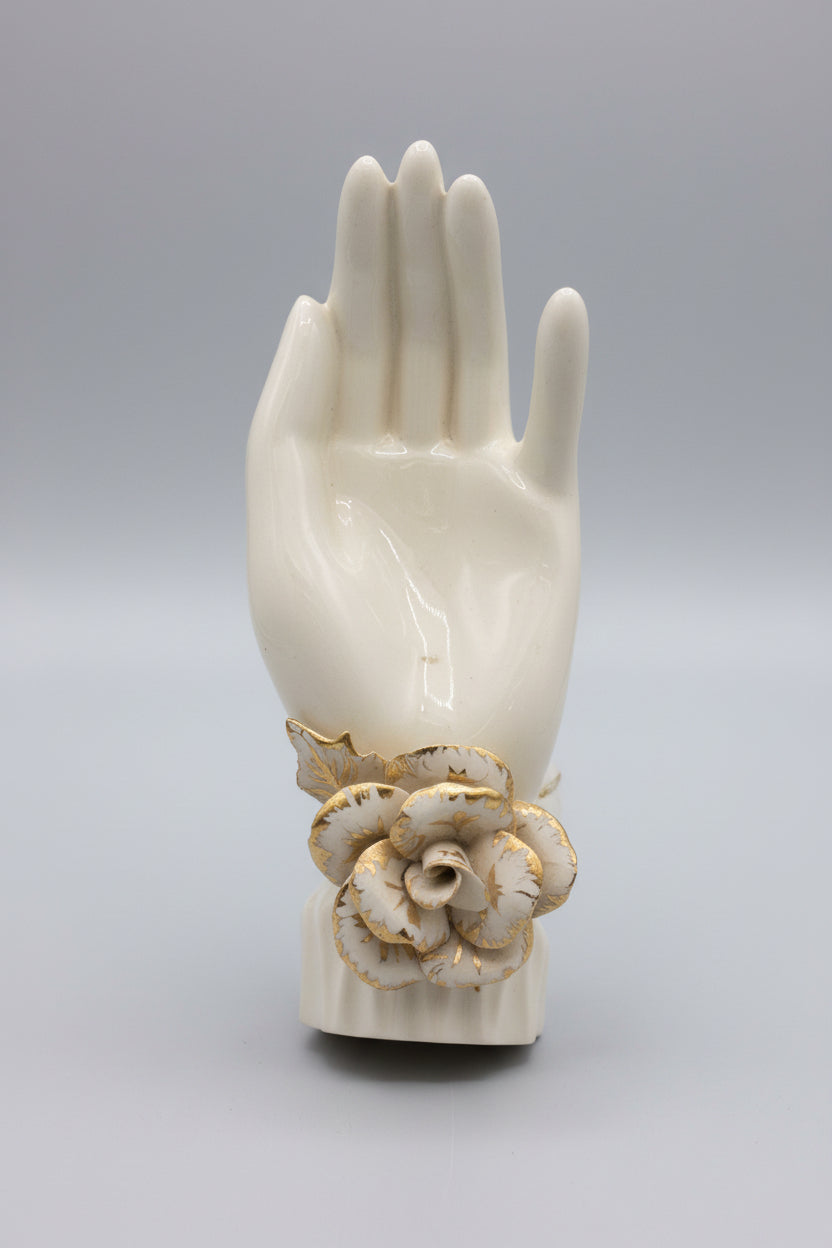 An off-white ceramic or resin wall-mounted hand sculpture holding a decorative cream and brown flower at the wrist, set against a dark brown paneled background.