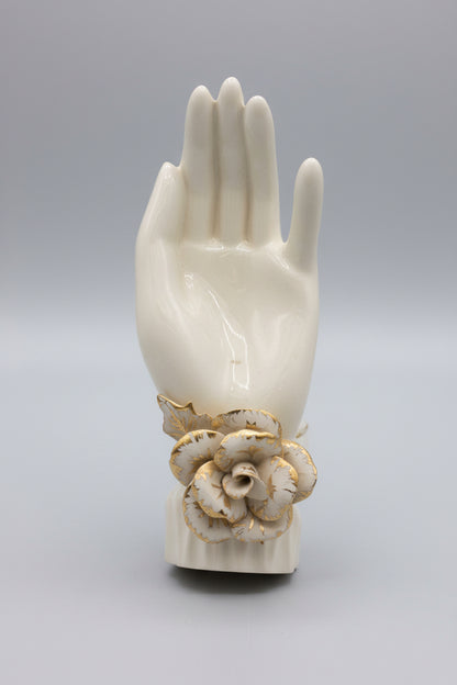 An off-white ceramic or resin wall-mounted hand sculpture holding a decorative cream and brown flower at the wrist, set against a dark brown paneled background.