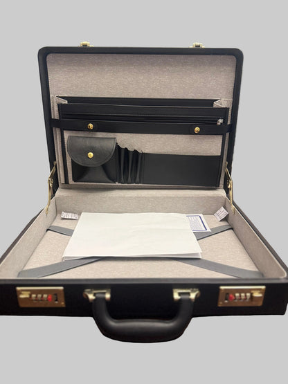 An open black briefcase-style organizer case on a wooden table, showing interior pockets, a gray strap across the base, and a white paper insert.