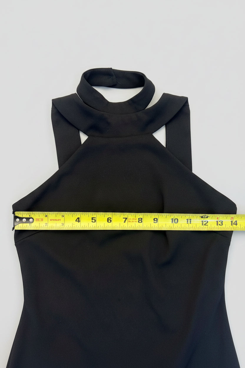 A black sleeveless halter-neck top laid flat on a wooden floor, with a yellow tape measure stretched horizontally across the chest to show the width.