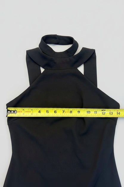 A black sleeveless halter-neck top laid flat on a wooden floor, with a yellow tape measure stretched horizontally across the chest to show the width.