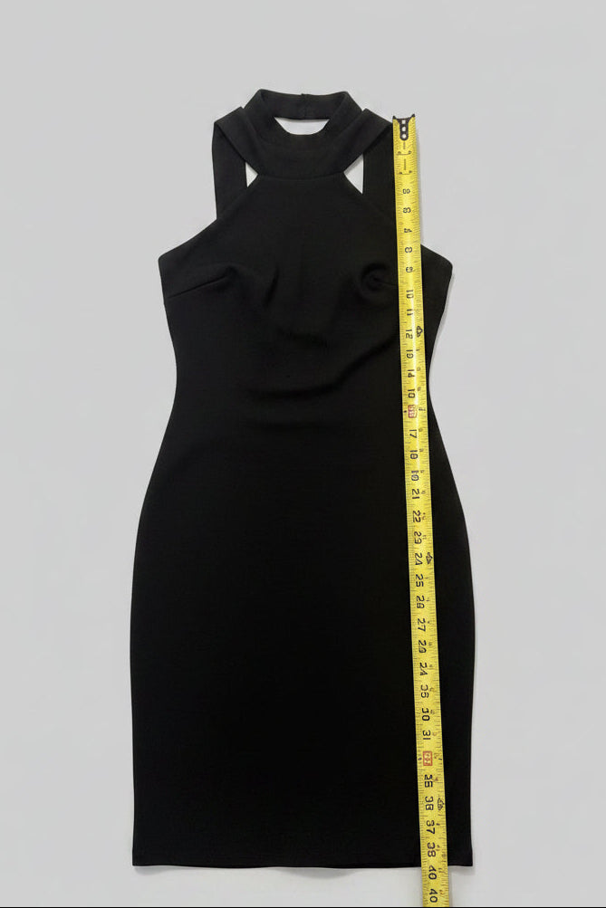 A sleeveless black dress laid flat on a wooden table next to a yellow measuring tape, with various household items visible around it.