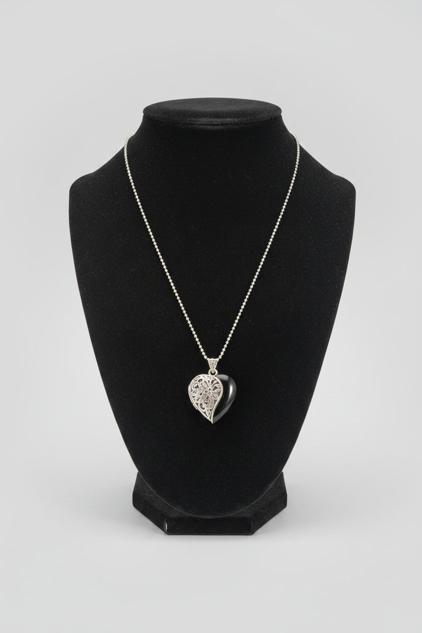 A silver heart-shaped pendant necklace with an engraved pattern, displayed on a black velvet jewelry bust over a wooden table.
