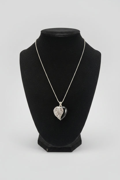 A silver heart-shaped pendant necklace with an engraved pattern, displayed on a black velvet jewelry bust over a wooden table.