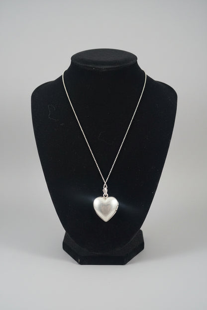 A silver heart-shaped pendant necklace displayed on a black jewelry bust over a wooden surface.