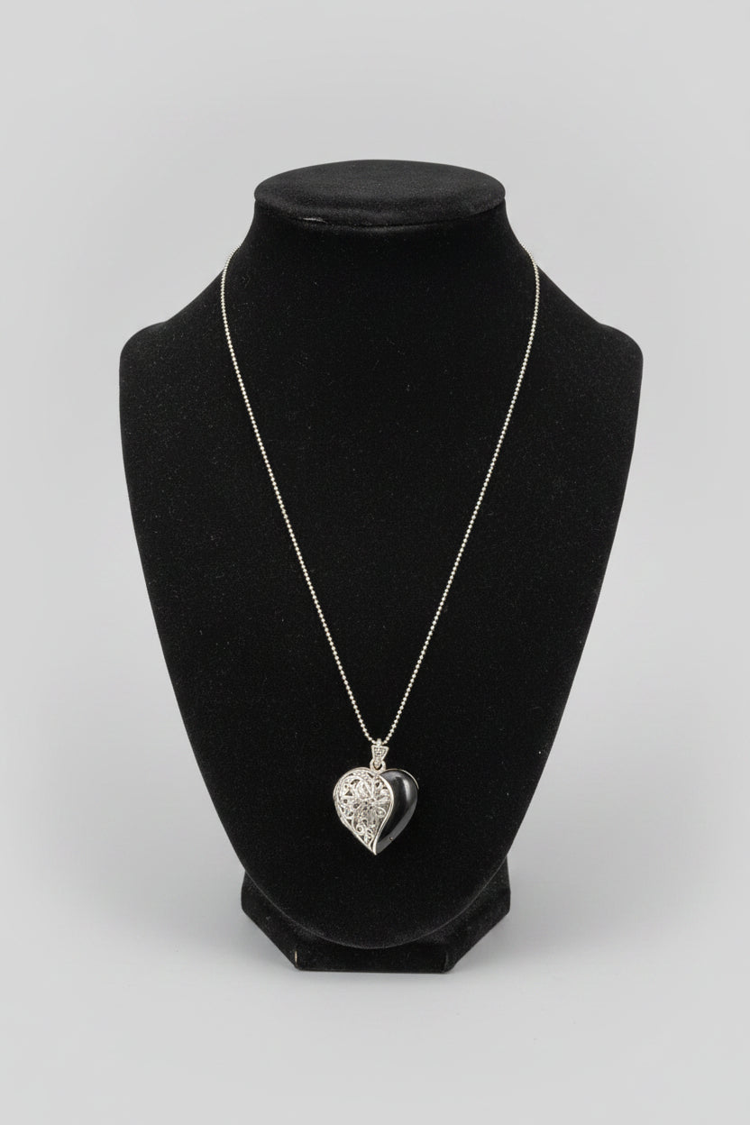 A silver heart-shaped pendant necklace displayed on a black jewelry bust, set on a wooden table with a blurred indoor background.