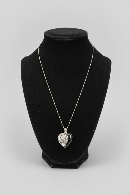 A silver heart-shaped pendant necklace displayed on a black jewelry bust, set on a wooden table with a blurred indoor background.