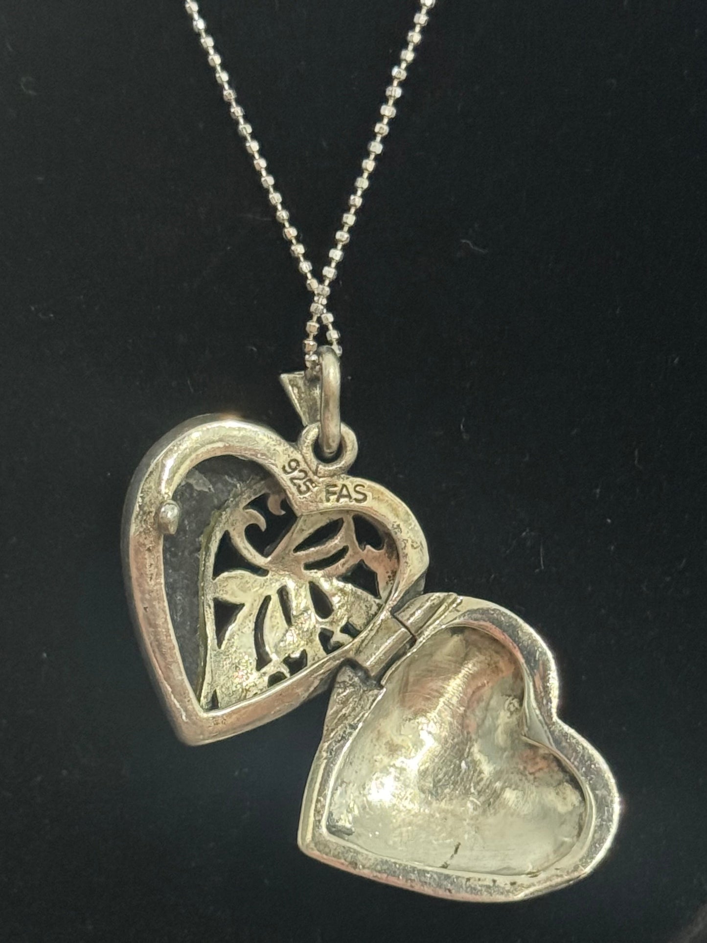 A silver heart-shaped locket pendant necklace on a matching chain, displayed on a black jewelry bust. The locket is open, showing one side with a floral cutout and the other with a textured, polished finish.