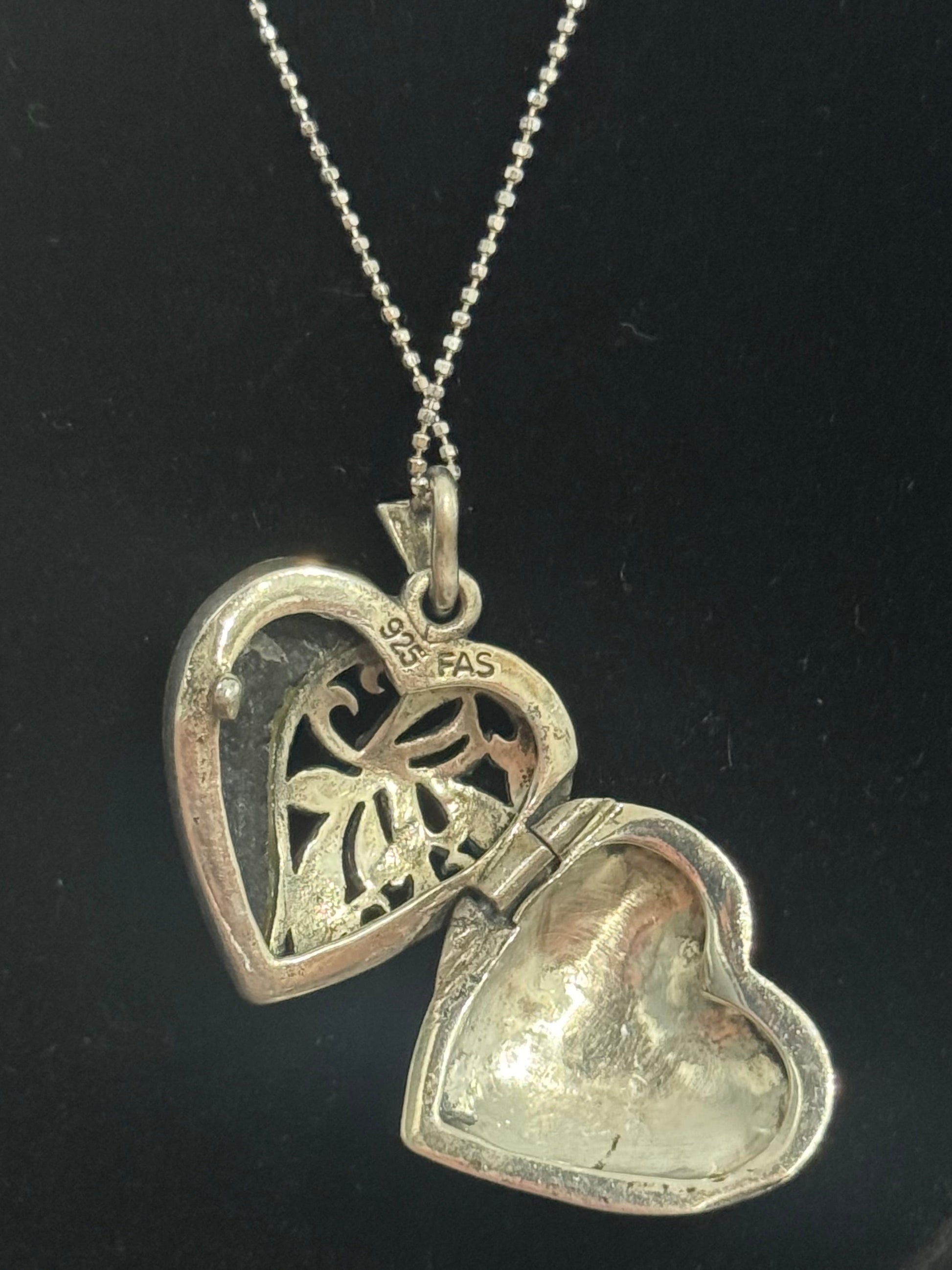 A silver heart-shaped locket pendant necklace on a matching chain, displayed on a black jewelry bust. The locket is open, showing one side with a floral cutout and the other with a textured, polished finish.