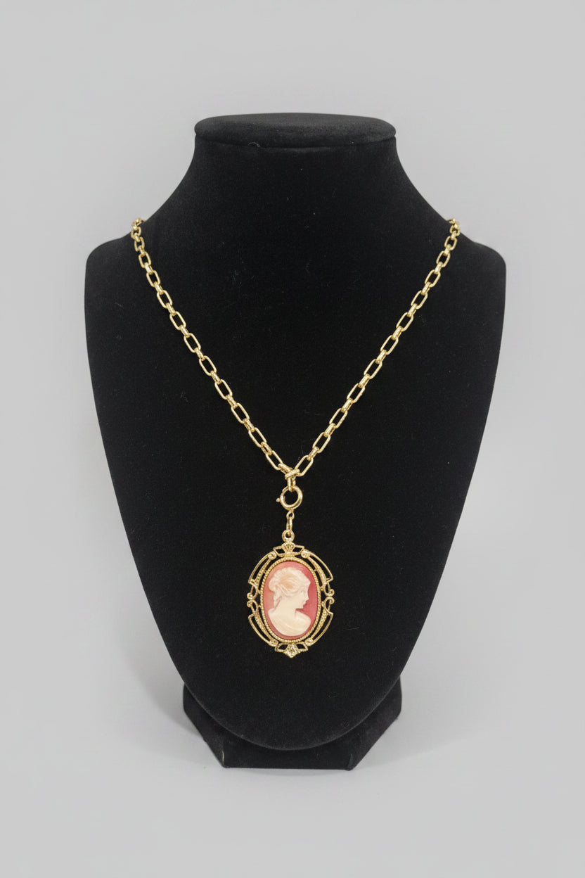 A gold chain necklace with an oval pendant featuring a pink and cream cameo portrait, displayed on a black velvet jewelry bust.