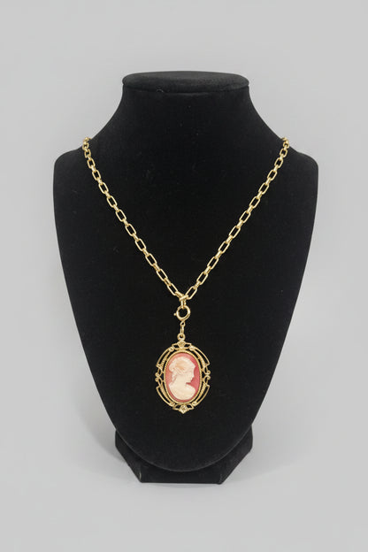 A gold chain necklace with an oval pendant featuring a pink and cream cameo portrait, displayed on a black velvet jewelry bust.
