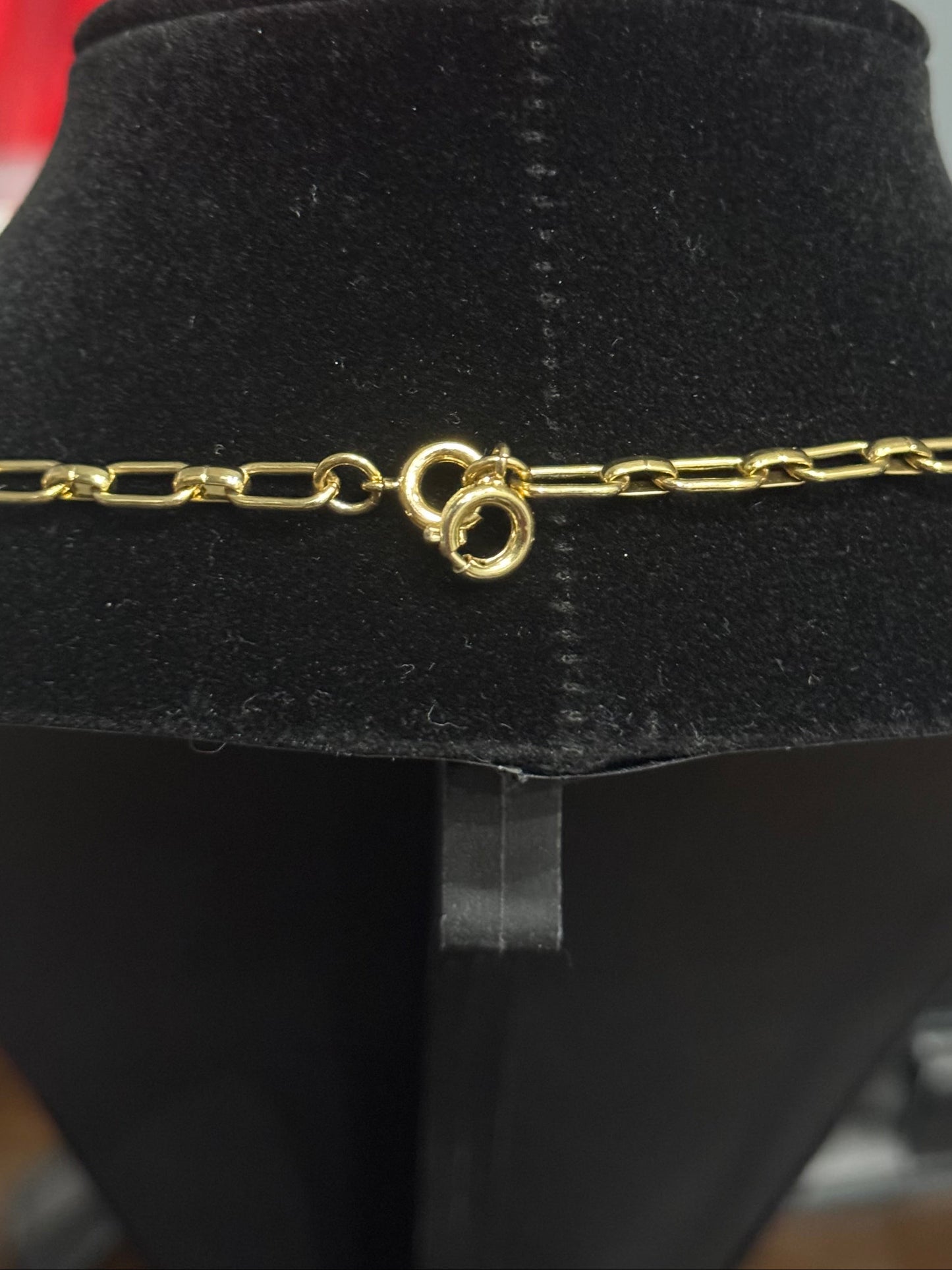 A gold chain necklace displayed on a black velvet jewelry bust, showing a lobster clasp closure. The background is a blurry indoor setting with a person visible in the upper area.