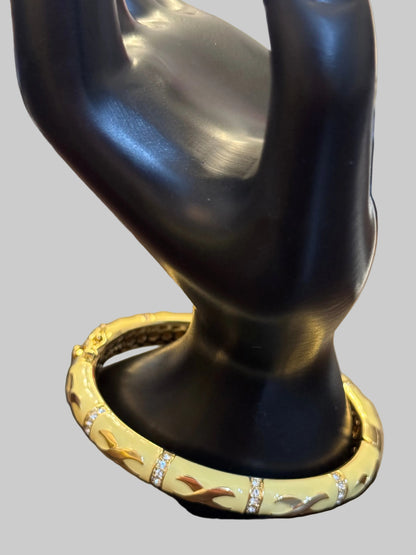 A gold bangle bracelet with a textured pattern and small clear stone accents, displayed on a black jewelry stand over a wooden table.