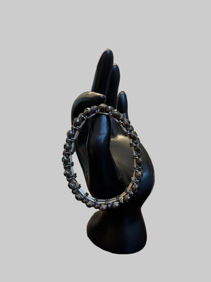 A silver-toned chain-link ring displayed on a black mannequin hand, set on a wooden table with a blurred jewelry store background.