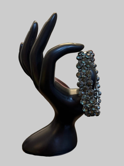 A large, wide silver ring encrusted with sparkling clear stones, displayed on a black hand-shaped jewelry stand on a wooden table. The background shows a cluttered home environment with a clock, boxes, and other jewelry.