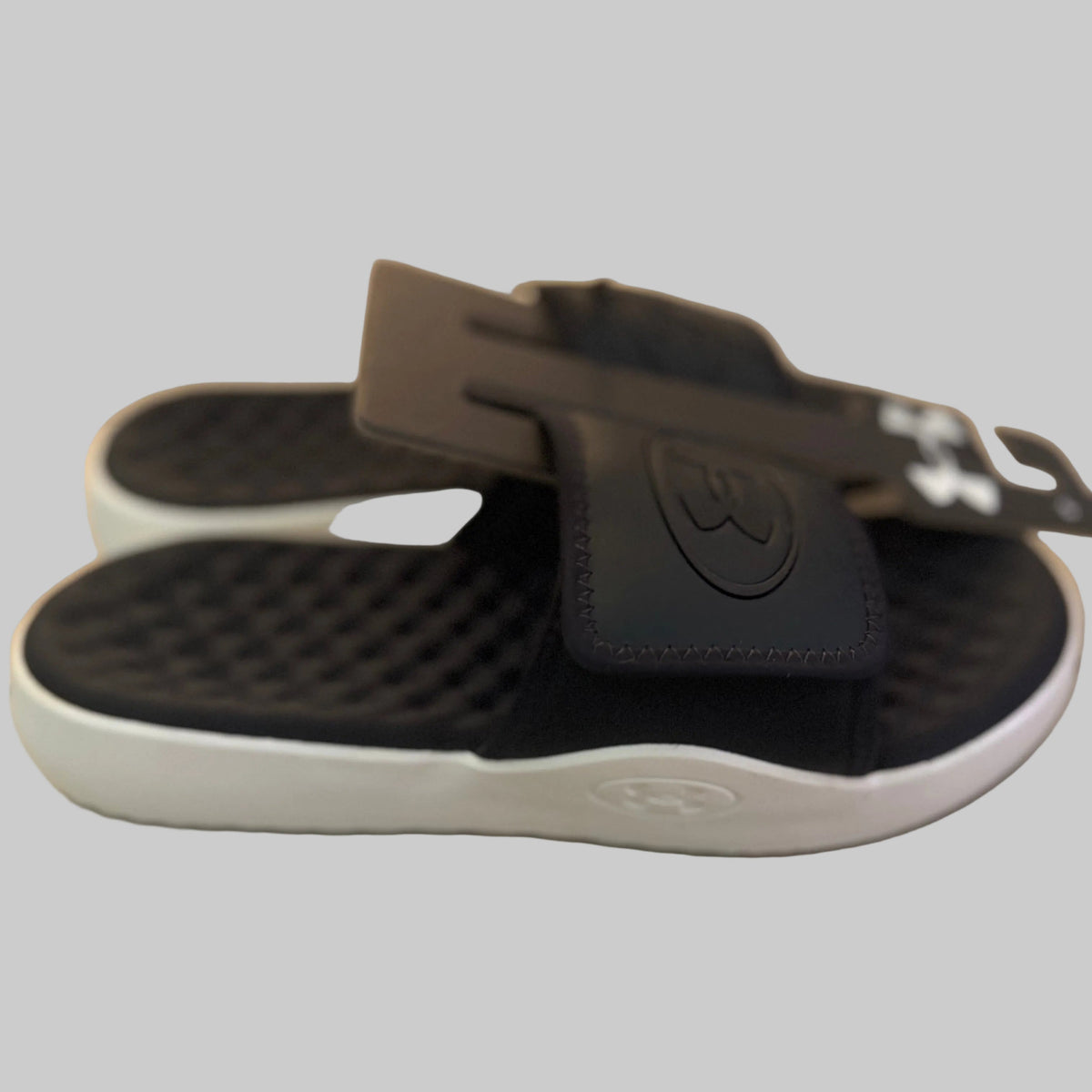 Black slide sandals with a white sole, featuring a textured footbed and a wide strap with a debossed logo, resting on a light-colored surface.