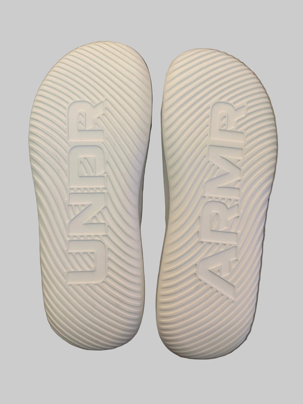 A pair of white athletic shoes shown from the sole side, featuring the embossed brand names 'UNDR' and 'ARMR' on the tread, resting on a light-colored wooden surface.