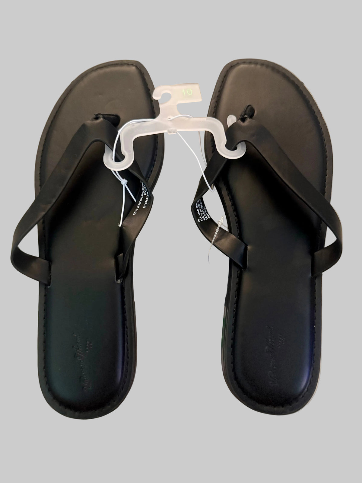 A pair of black leather thong sandals with flat soles, shown on a light-colored wooden floor. One sandal is on a clear plastic hanger, and the other has a paper tag attached.